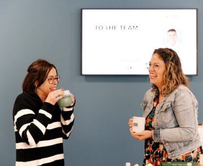 Deux femmes souriantes discutent autour d'une tasse, devant un écran affichant "TO THE TEAM