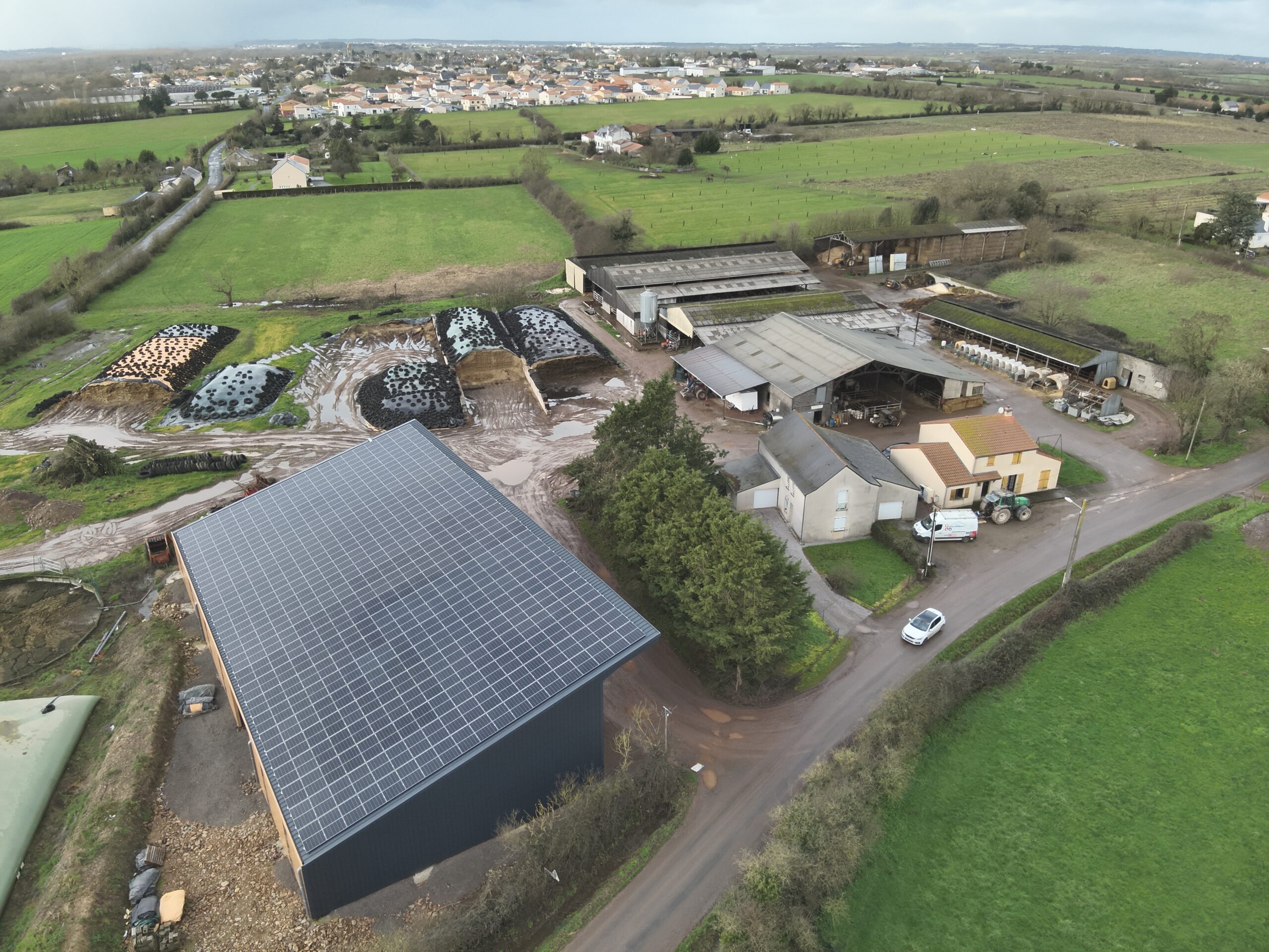Vue aérienne d'une ferme avec panneaux solaires, bâtiments agricoles et champs verts.